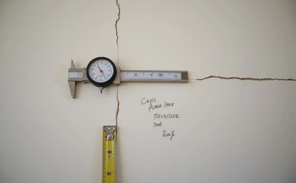 Crack gauge fixed across a diagonal wall crack with a dated reference mark and tape measure